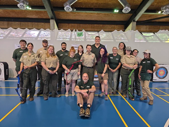 A group of people wearing uniforms are standing and kneeling indoors on a blue floor. Some hold archery bows. Targets can be seen in the background.