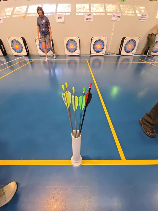 An indoor archery range with several targets is shown. Six arrows with different colored fletchings are standing in a holder. Several people are visible in the periphery of the image.
