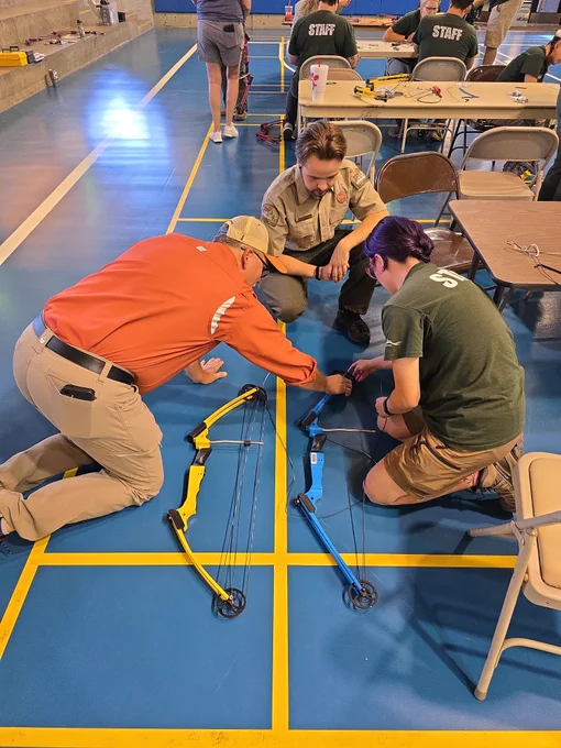 Three individuals are kneeling on a gym floor, assembling a blue and yellow compound bow. They are surrounded by several chairs and tables. Some chairs in the background have the word "STATE" printed on them.