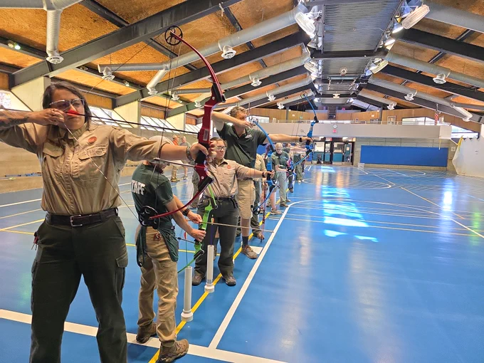 A group of individuals practicing archery inside a large sports hall with blue flooring and high ceilings.
