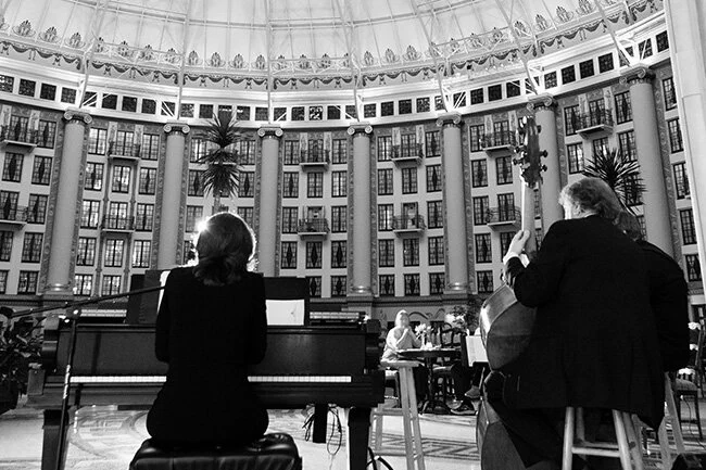 Two musicians perform inside West Baden Springs Hotel atrium with large windows. One plays a piano while the other plays a double bass. The setting conveys an elegant and formal atmosphere.