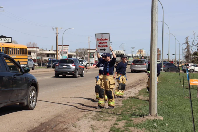 Edmonton Fire Rescue recruits stand by a busy street waving an orange traffic baton and holding a sign that reads "Firefighter Charity Carwash" to direct passing vehicles.