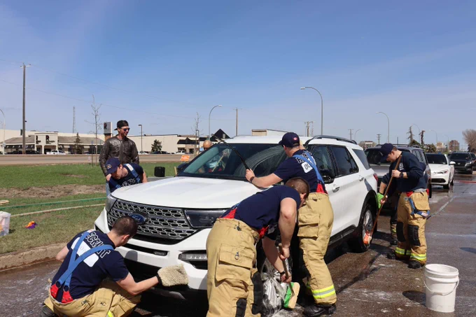 Rescue recruits in bunker pants and navy t-shirts work together to wash a white Ford SUV using brushes, mops, and mitts during a charity car wash.