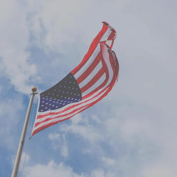 The image shows the United States flag waving on a pole against a backdrop of a partly cloudy sky.