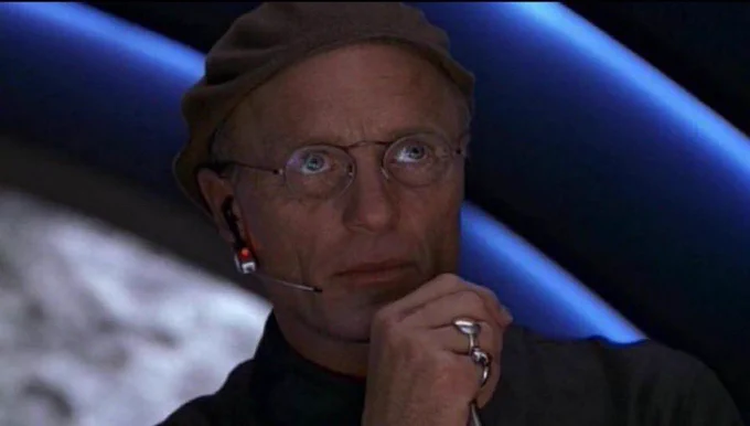 The Truman show christof scheming meme. Older man with glasses and a brown cap with headphone on his right ear, right hand held up to his chin with rings on two fingers