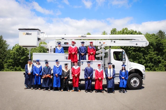 A group of FLEET graduates in red, blue, and navy caps and gowns posing in front of an Altec bucket truck, with some standing on the truck bed and others lined up on the ground below.