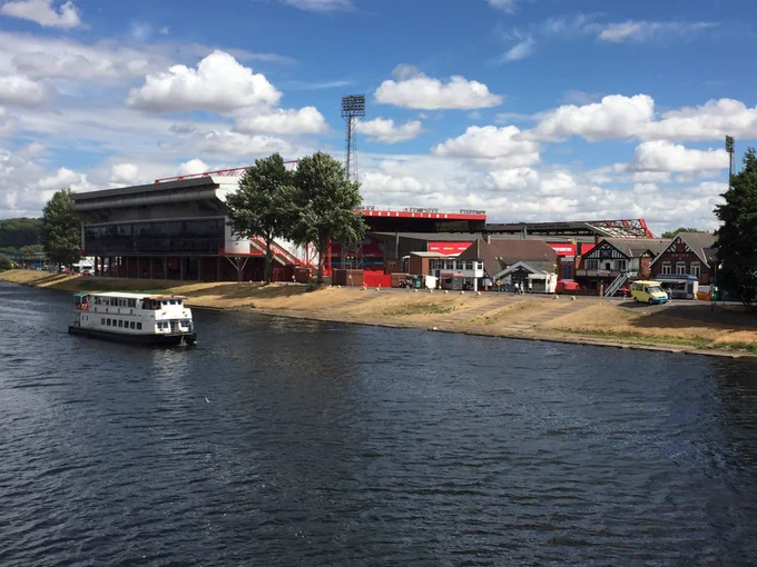 Photo of Nottingham Forest’s City Ground Stadium taken from Trent Bridge. The shot is taken in daylight hours, on a hot summer afternoon. Two thirds of the picture in the immediate foreground are the dark blue waters of the River Trent, upon which, a large white boat can be seen sailing away to the left. The Trent End stand is closest, with its huge white paneling angled towards the river. Two floodlights can be seen behind large bushy trees and the ground appears to be empty. The sky above the stadium is light blue interspersed with fluffy white clouds.