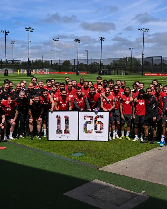 A photo showing Mo Salah and Andy Robertson posing with the LFC squad and staff, alongside framed 11 and 26 graphics. 