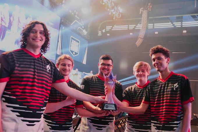 Five esports players in matching black and red jerseys hold a trophy together on a brightly lit stage.