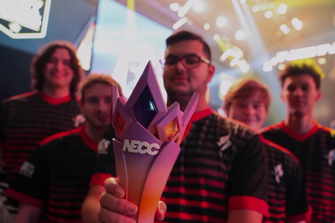 A team of esports players in matching black and red jerseys proudly holding a NECC championship trophy on a brightly lit stage.