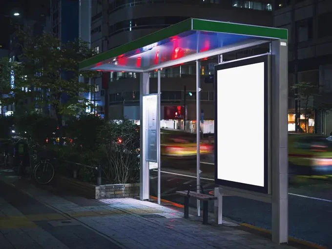 An illuminated bus shelter with a blank advertising panel on a city street at night.