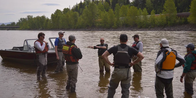 A group of people wearing life jackets are standing in a river near a boat. They appear to be listening to someone who is gesturing while speaking. Trees line the riverbank in the background.