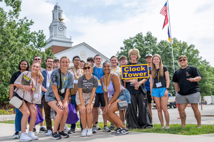 A group of people smiling and posing outdoors near a campus building with a clock tower and a flagpole displaying two flags. One person holds a sign that reads "Clock Tower."
