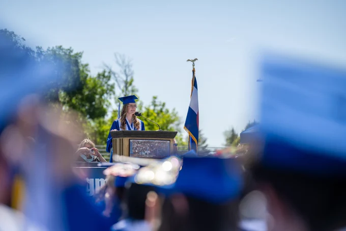 A female graduate in glasses stands at the podium and gives a speech to her classmates during a sunny outdoor graduation ceremony