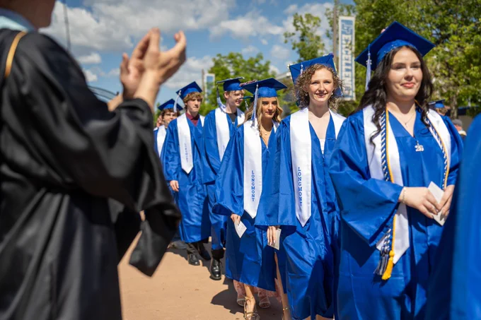 A line of graduates marches down a walkway in regalia, applauded by their teachers on a sunny day. 