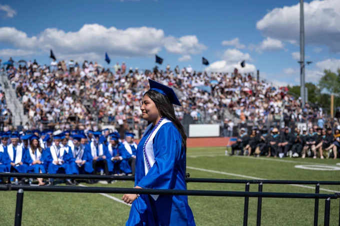 A graduate in a blue cap and gown walks across the field during an outdoor graduation ceremony, with classmates seated nearby and a crowd of family and friends in the stands behind.