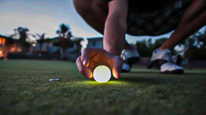 Close-up photograph of a person reaching down to pick up a glowing golf ball.
