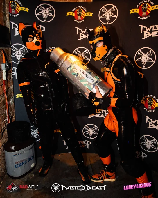 Elio in latex catsuit looks concerned as GimpSimp in orange K9 suit offers a large model bottle of poppers