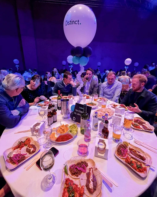 Round dinner table with guests, shared dishes, drinks, and a “Distinct.” branded balloon centrepiece at an event.