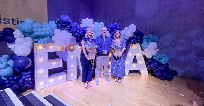 Three attendees posing with bouquets in front of lit letters and blue, white, and black balloon backdrop.