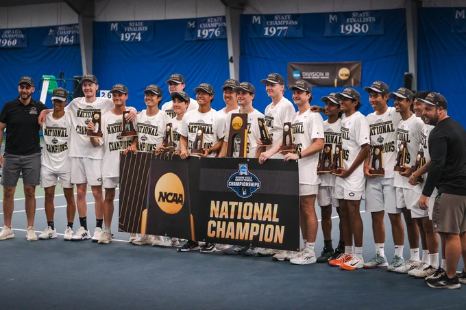 A team of male athletes and coaches pose indoors. They hold trophies and a banner reading \"2026 DIII MEN'S TENNIS CHAMPIONSHIPS, NATIONAL CHAMPION\". Banners in background mention STATE FINALISTS in 1974, 1976, 1977, and 1980.