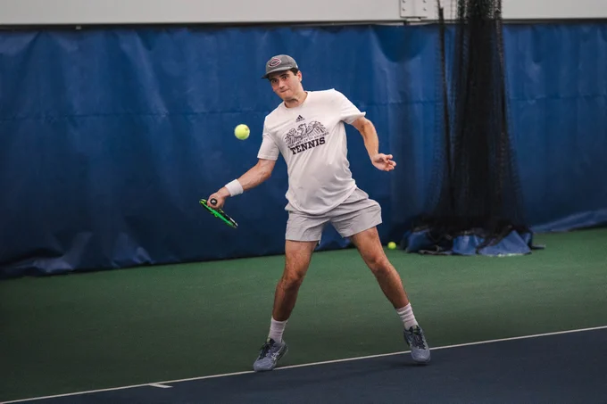 A tennis player in a hat and \"THE UNIVERSITY OF CHICAGO TENNIS\" shirt swings at a ball on an indoor court. The player wears gray shorts and athletic shoes.