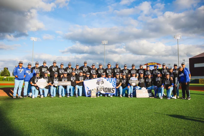 Wide team photo taken on a baseball field. A large group of players and staff wear matching black “2026 Summit League Baseball Champs” shirts, light blue pants, and championship hats with medals around their necks. The group is arranged in multiple rows, with some kneeling in front and others standing behind. Several individuals hold championship plaques and a large banner reading “2026 Baseball Champions". 