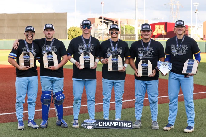 Six baseball players stand side by side on a baseball field, wearing matching black “2026 Summit League Champs Baseball” shirts, light blue pants, and championship hats. Each player has a medal around the neck and holds an award plaque labeled “Summit League All-Tournament Baseball 2026,” while one player holds an additional plaque marked “MVP.”