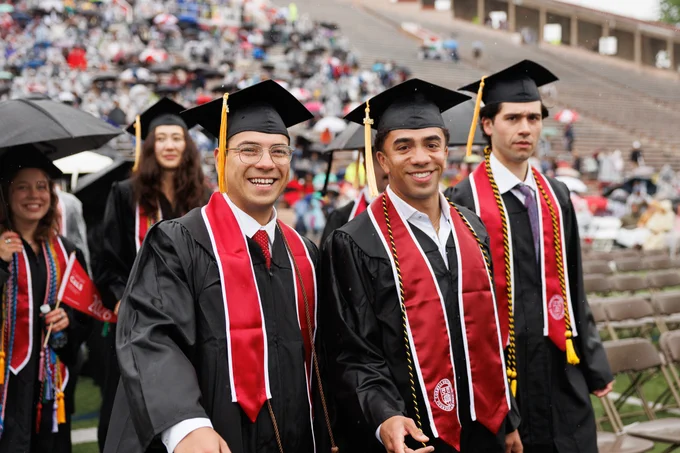 A group of Cornell graduates in black caps and gowns with red and white sashes stand together smiling at an outdoor ceremony. The background shows a stadium with many people seated and some holding umbrellas.