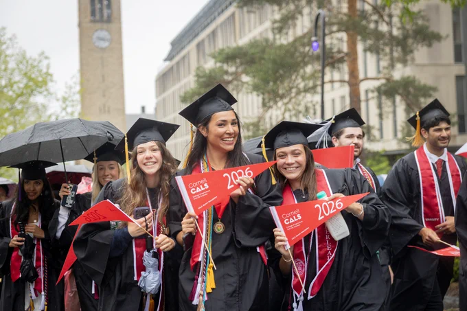 A group of people in graduation attire are smiling and holding red pennants with "Cornell Class of 2026" printed on them. One person holds an umbrella. In the background, a building and a clock tower are visible.