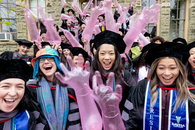 A group of people in graduation attire, including caps and gowns, are celebrating outdoors. Some are holding inflated pink gloves, and there is a mix of joyful expressions. A diverse range of clothing and sashes is visible. The setting is in front of a stone building.