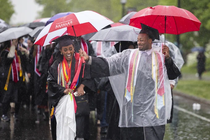 Cornell Graduates walking in the rain, wearing caps and gowns with colorful umbrellas. Some hold umbrellas with Cornell branding. The atmosphere is joyful despite the wet weather.