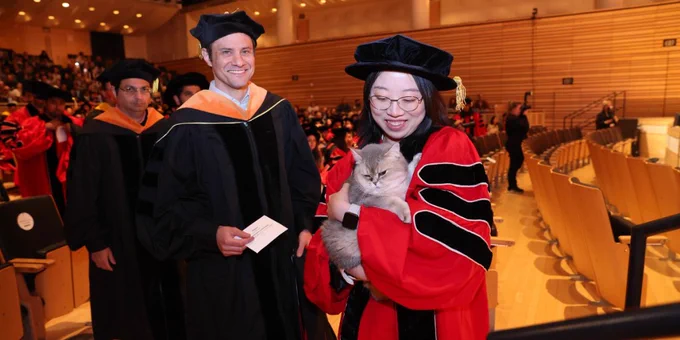 A person in academic regalia holds a fluffy cat at a graduation ceremony. Another person stands nearby, smiling, also in academic attire. The setting is an auditorium filled with people.