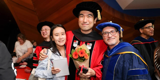 A graduate in academic regalia holds a bouquet and poses with two others at a ceremony. People in similar attire are visible in the background.