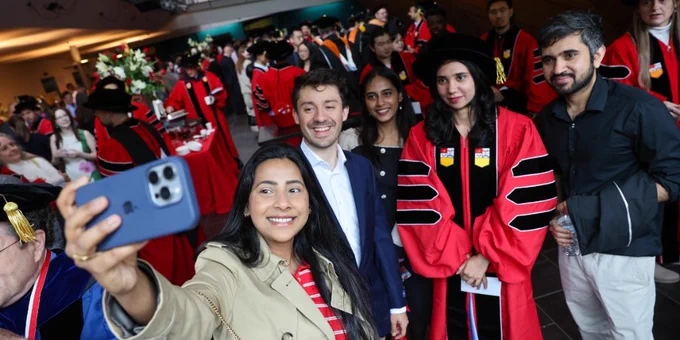 A group of individuals is posing for a selfie at a graduation event. Many are wearing academic regalia, including red gowns and caps with gold tassels. The setting is a crowded indoor venue with people in the background.