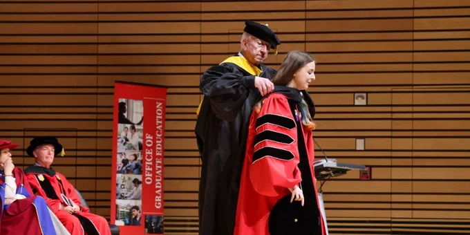An academic ceremony where a person is being awarded a doctoral hood on stage. Others in academic regalia are seated nearby. A banner reads "Office of Graduate Education.