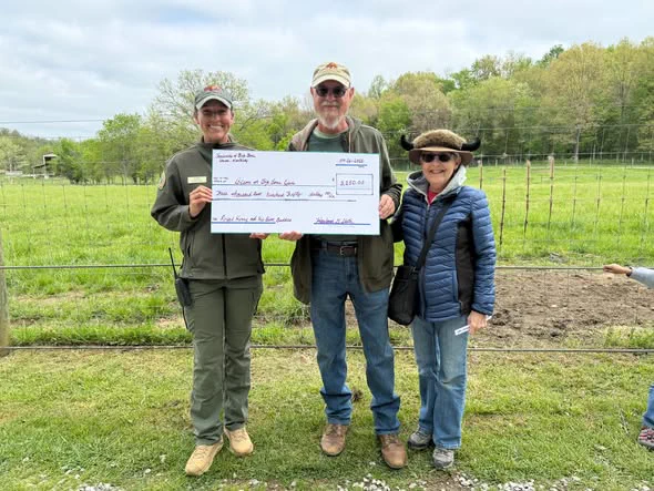 Three people stand in a grassy field holding a large check, with one person holding a camera. The check indicates a sum of $3,000, made out to "Citizens for Big Sky Park." There are trees and a fence in the background.