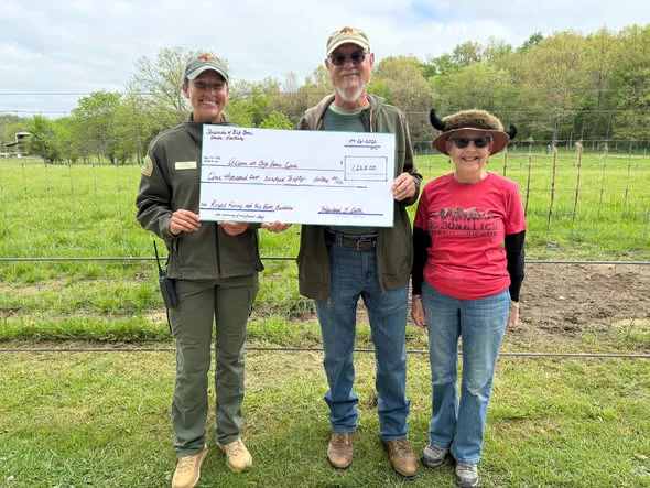Three individuals stand outdoors holding a large ceremonial check for $1,868.00 from the Friends of the Blue Ridge Parkway, addressing the Roanoke Star Sky Camp. They are in a grassy area with trees in the background.