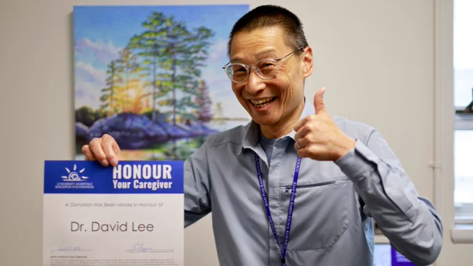 Dr. David Lee smiling in his office holding his Honour Your Caregiver certificate in one hand while giving a thumbs up with the other.