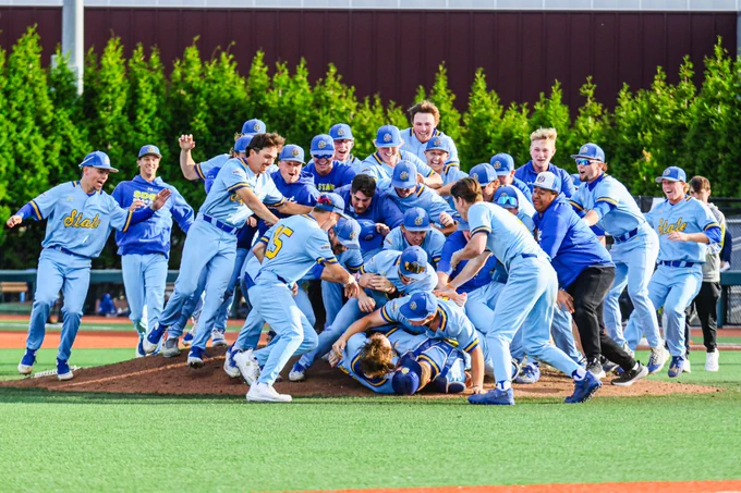 Several baseball players in light blue uniforms rush toward the mound, where more teammates embrace at the center. Surrounding players run in from all directions with arms outstretched, preparing to join the celebration. The infield dirt and green outfield grass are visible, with a fence and spectators in the background