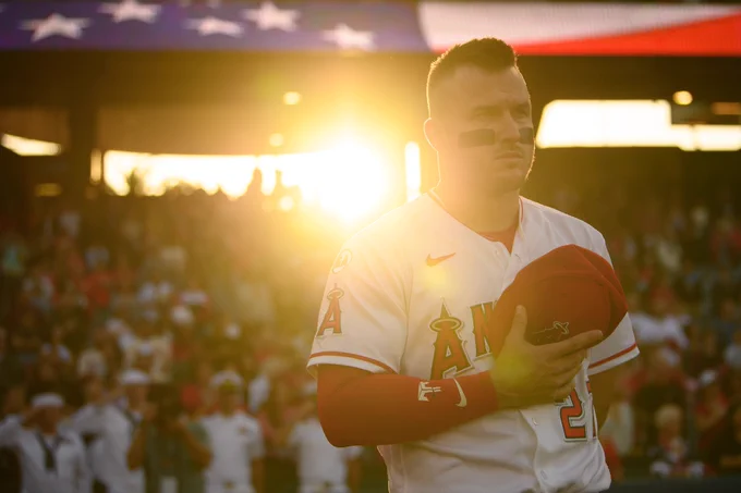 Mike Trout is backlit by the setting sun as he stands with his hat over his heart during the national anthem.