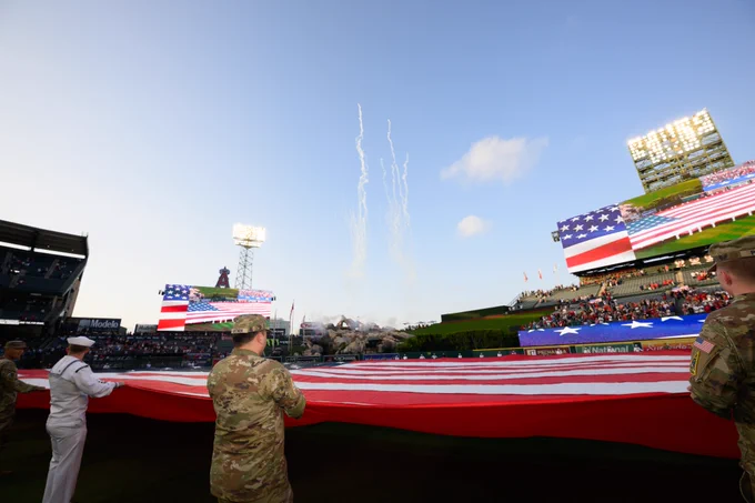 Fireworks shoot off in the background while service members hold a large outstretched flag across the outfield at Angel Stadium.