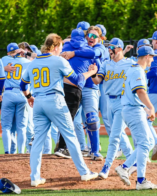 On the pitcher’s mound, a group of baseball players in light blue uniforms gathers in celebration.
