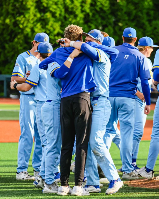 On the pitcher’s mound, a group of baseball players in light blue uniforms gathers in celebration.