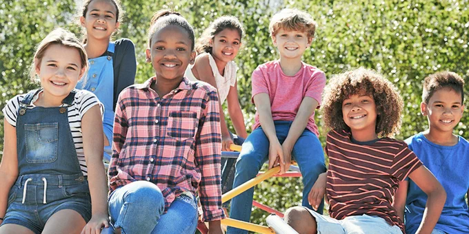 Group of seven diverse children sitting and standing outdoors on playground equipment on a sunny day.