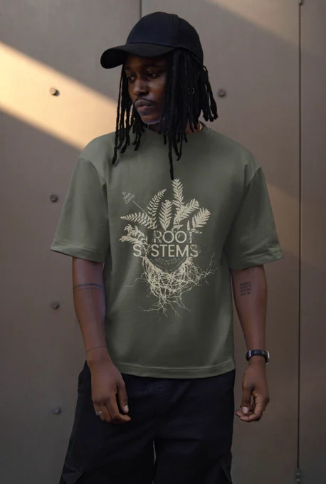 A person wearing the DWeb Camp 2026 T-shirt. Its colour is green khaki, and it has a light overlay of foliage with the text “Root Systems”