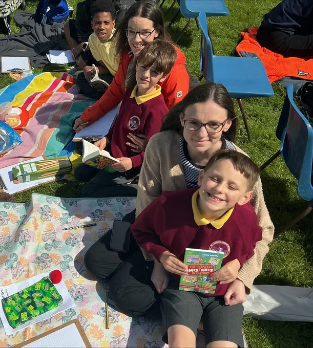 Children and adults in school uniforms reading books outdoors on a sunny day.