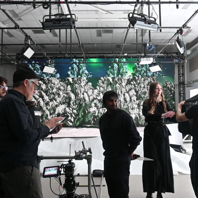 Film crew and actors collaborate on a studio set with professional lighting and camera equipment capturing a scene against a snowy forest backdrop.