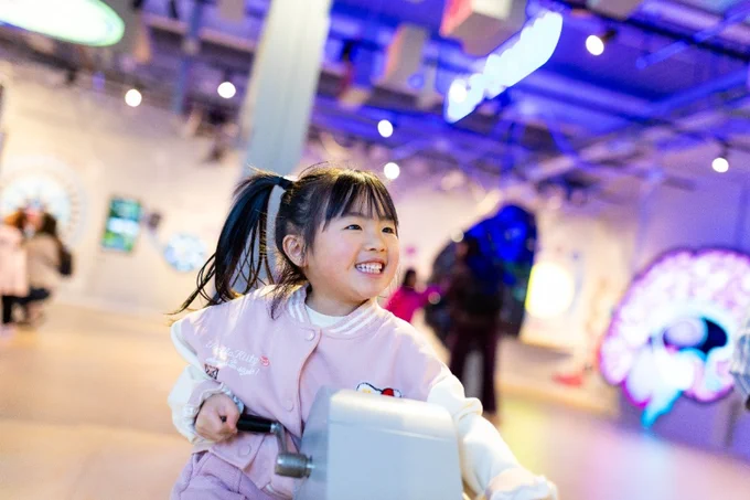 Young girl with a ponytail riding a stationary bike in a brightly lit indoor area.