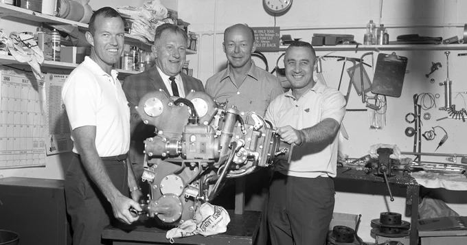 Gus Grissom and three others are standing in a cluttered workshop, gathered around a complex mechanical device on a table. The walls are lined with various tools and equipment.
